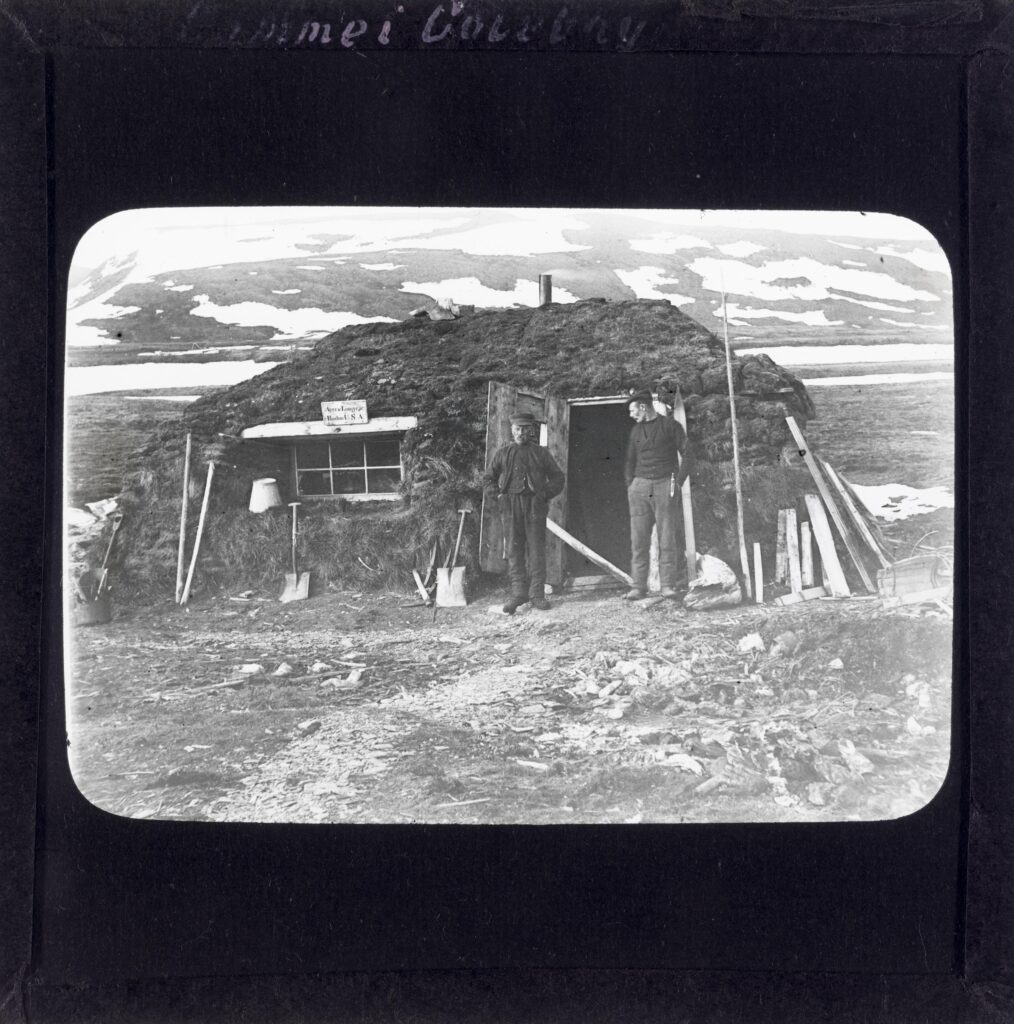 Slide belonging to dr. philos. Gunnar Holmsen, used in lectures, teaching, etc. Gamme, or peat hut used as a hunting cabin in 1908 in Colesbukta, Svalbard. Two men stand outside the hut. The sign on the hut reads "Ayer and Longyear Boston USA.", i.e. the company Ayer & Longyear, which bought the Trondhjem-Spitsbergen Kulkompani company in 1904. The men are not identified. 